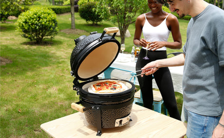 Gril en céramique Kamado noir, avec pizza garnie, utilisé par un homme et une femme dans un jardin verdoyant et ensoleillé