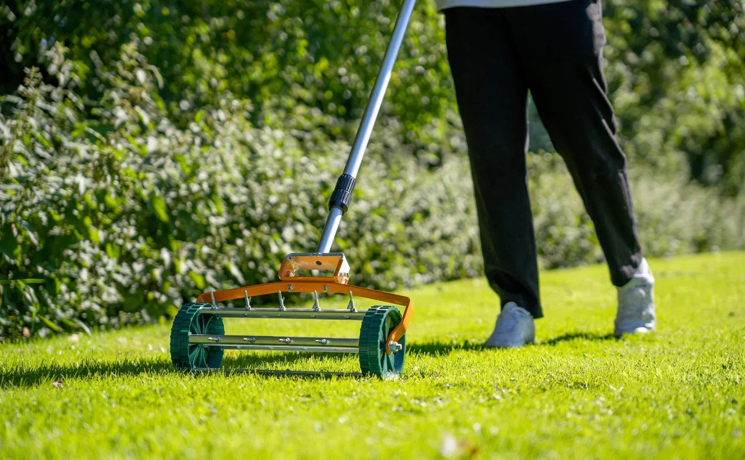 Un épandeur à gazon orange et vert en action sur une pelouse verte, avec une personne en pantalon noir et chaussures blanches en arrière-plan