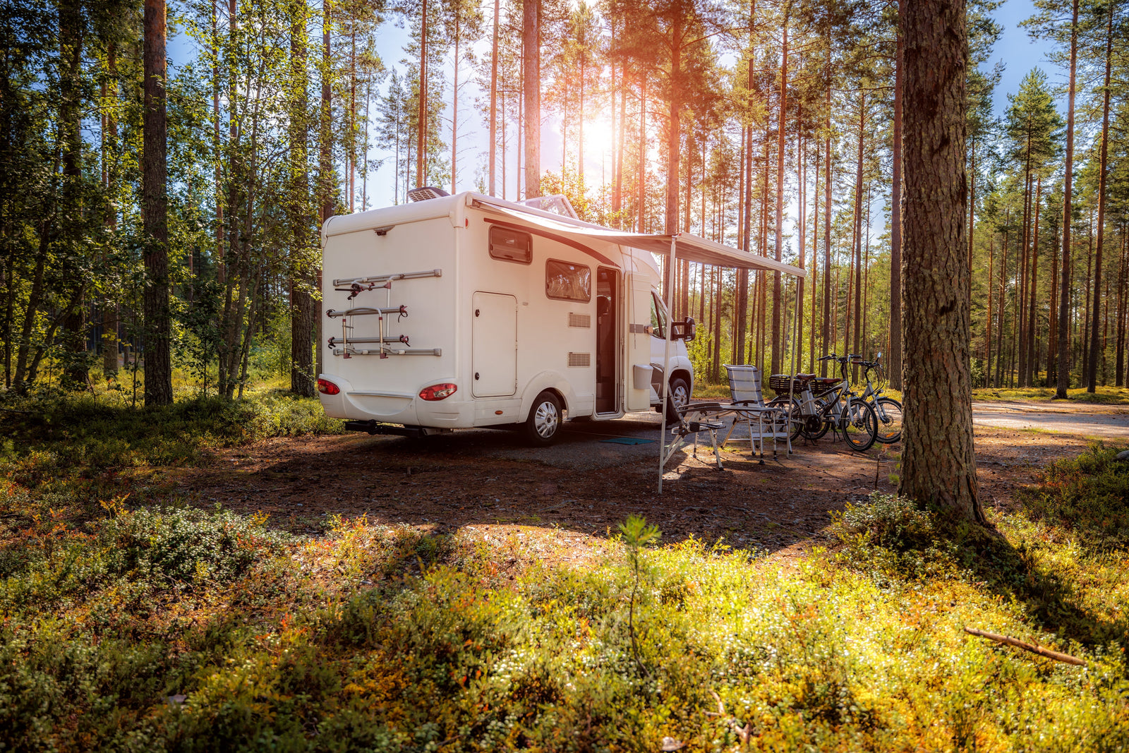 Camping-car blanc stationné dans une forêt verdoyante, avec des chaises pliantes et des vélos à côté, sous un ciel ensoleillé
