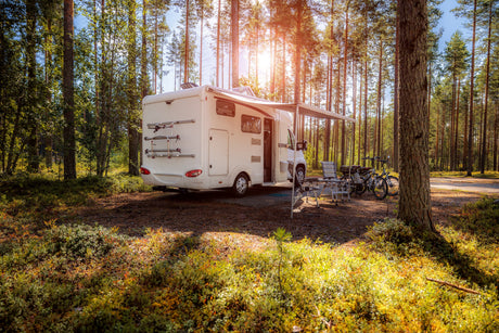 Camping-car blanc stationné dans une forêt verdoyante, avec des chaises pliantes et des vélos à côté, sous un ciel ensoleillé