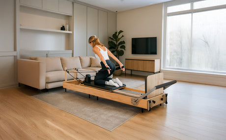 Femme en tenue de sport utilisant un reformer Pilates en bois clair dans un salon moderne avec un canapé et une grande fenêtre