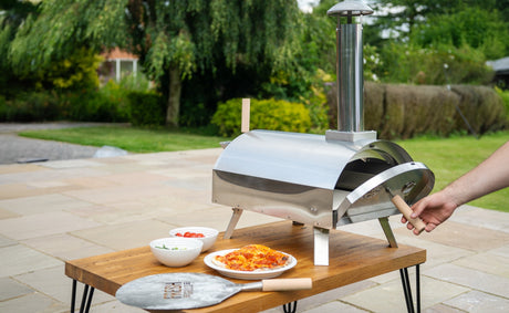 Un four à pizza en acier inoxydable sur une table en bois, avec des plats de tomates et de salade, prêt pour un barbecue d'été en plein air