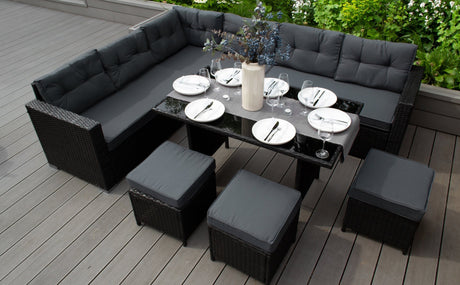 Salon de jardin en rotin noir avec coussins gris, table en verre, assiettes blanches et décor floral, sur une terrasse en bois