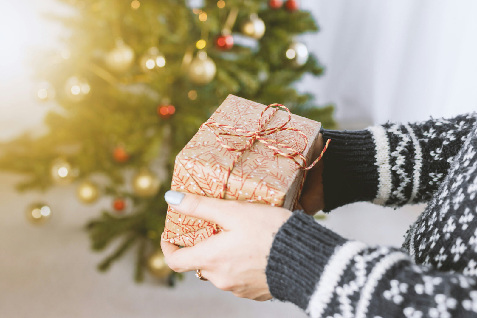 Cadeau joliment emballé avec un papier doré et un ruban rouge, tenu par des mains devant un sapin de Noël décoré