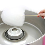 Barbe à papa blanche en train d'être enroulée sur un bâton, avec une machine à barbe à papa en métal brillant en arrière-plan