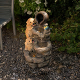 Fontaine de jardin en pierre avec trois chiots en résine, un pot en terre cuite et un éclairage LED, entourée de verdure