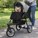 Poussette jogger pour animal de compagnie grise avec un petit chien à l'intérieur, roues noires et cadre léger sur un chemin verdoyant