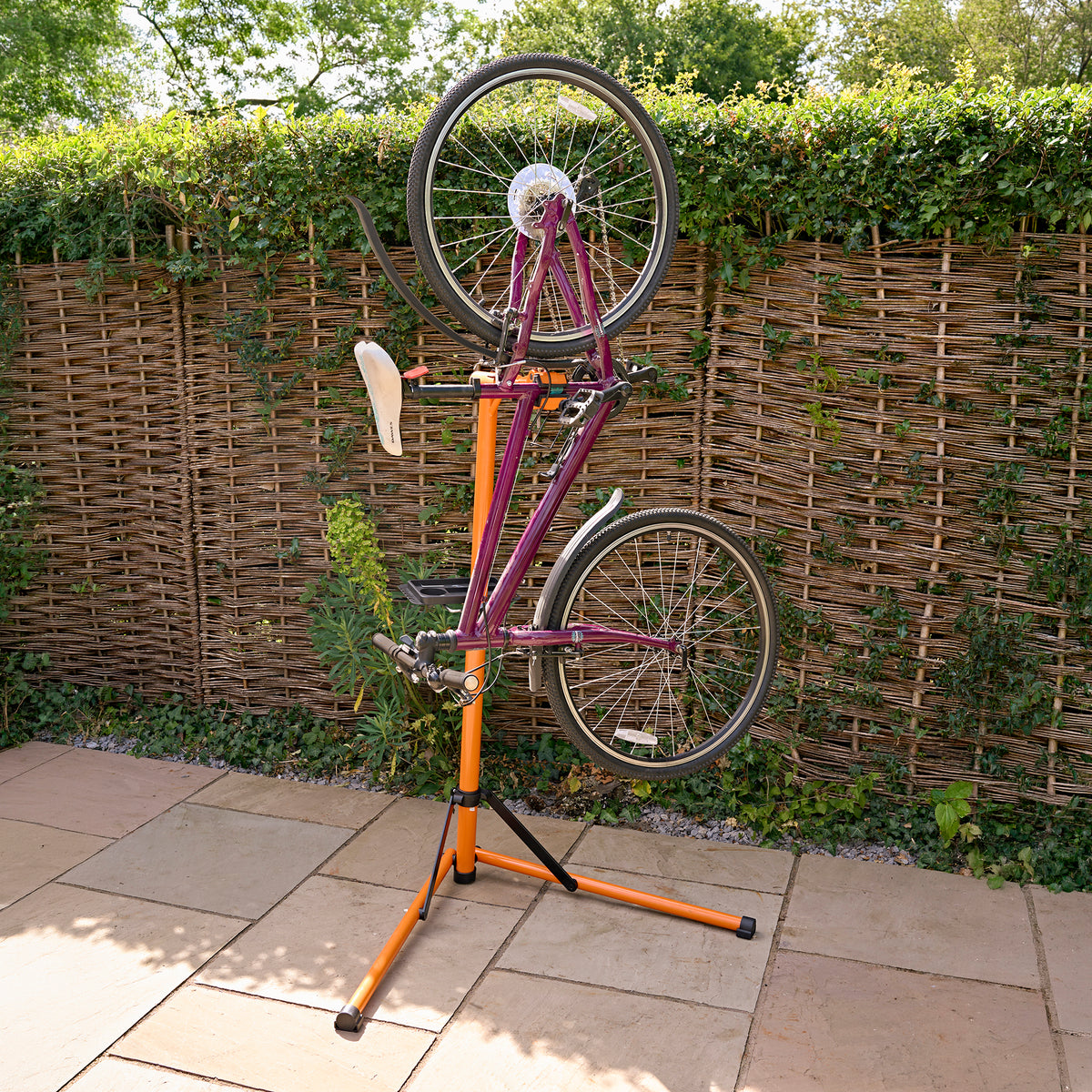 Pieds d'atelier mécanique Deluxe pour vélo, support orange et noir, tenant un vélo rose à l'envers sur un fond de haie verte