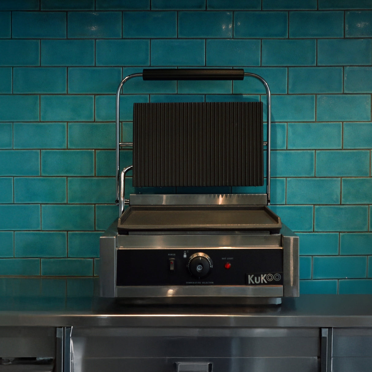 Presse à panini en acier inoxydable avec plaque rainurée, posée sur un comptoir, devant un mur carrelé turquoise brillant