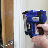 Agrafeuse-cloueuse électrique T-Mech bleue, tenue à la main, en train de fixer des clous sur un mur blanc près d'une porte en bois