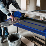 Scie de carrelage sur table, couleur bleue, avec un utilisateur plaçant une grande tuile grise sur la surface de coupe en métal