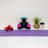 Haltères colorés sur un support noir, avec un tapis de yoga violet et des plantes en pot, créant un espace d'entraînement accueillant