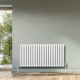 Radiateur à colonnes ovales blanc, 600 x 1200 mm, installé sur un mur gris, avec un sol en bois clair et une vue sur l'extérieur verdoyant