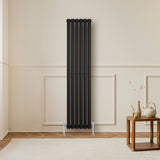 Radiateur à colonnes ovales noir, 1600 x 360 mm, installé sur un mur clair, avec un sol en bois et une table en bois à proximité