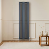 Radiateur à colonnes ovales gris anthracite, 1800 x 480 mm, installé sur un mur clair, avec un sol en bois et une table en bois à côté