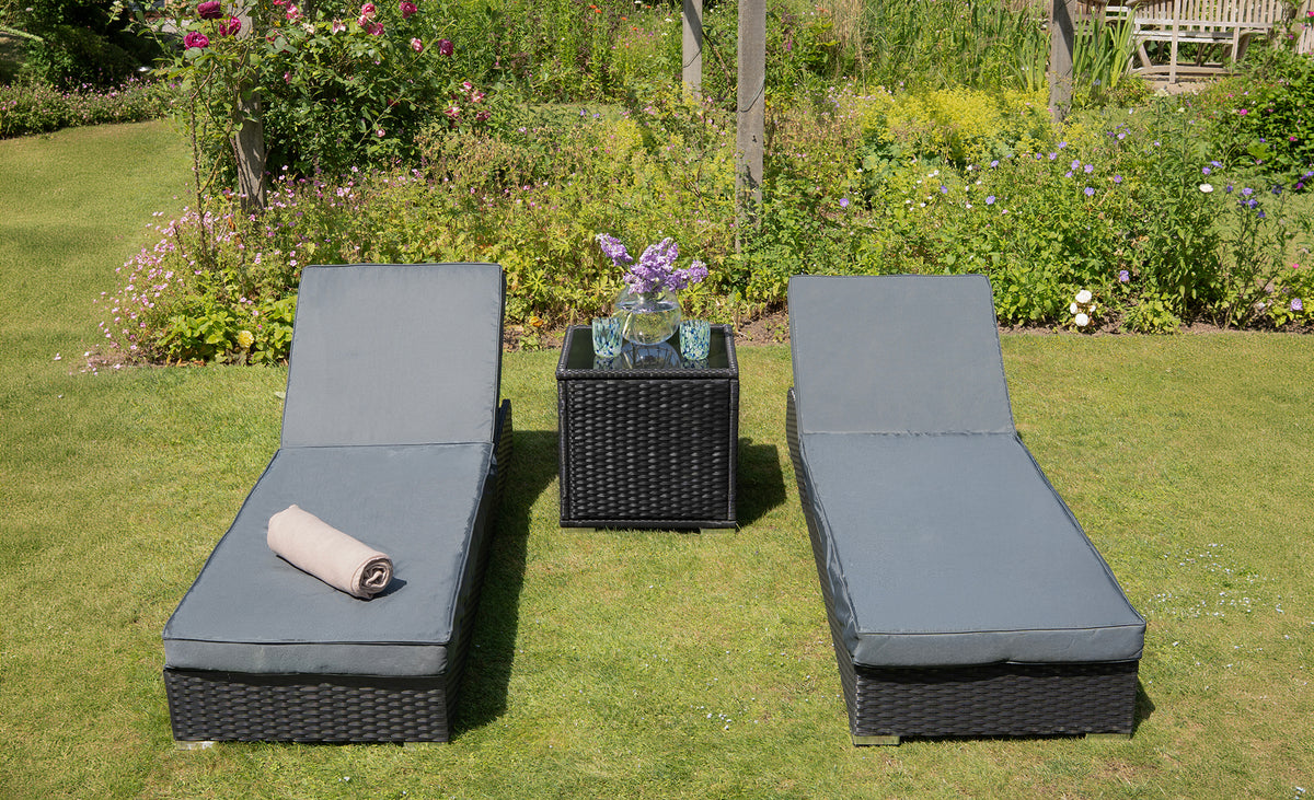 Chaises longues en osier noir avec coussins gris, serviette roulée, table basse en osier, entourées de fleurs colorées dans un jardin verdoyant