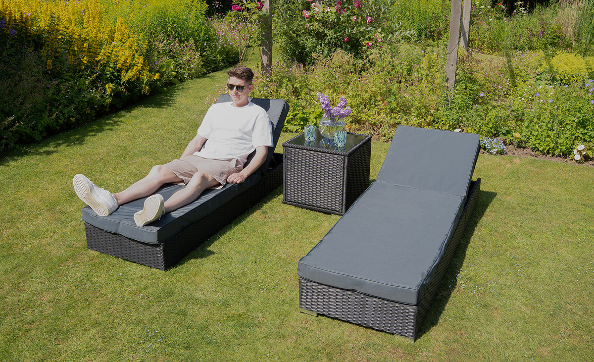 Un homme détendu sur un transat gris en osier, entouré de verdure et de fleurs, avec une table basse en osier à côté