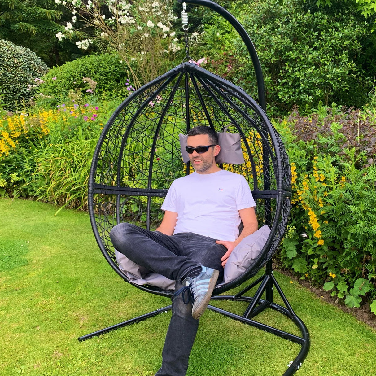 Chaise suspendue en rotin gris avec coussins, forme en goutte, installée dans un jardin verdoyant, offrant confort et style