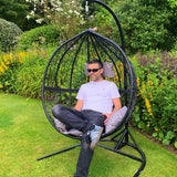 Chaise suspendue en rotin gris avec coussins, forme en goutte, installée dans un jardin verdoyant, offrant confort et style