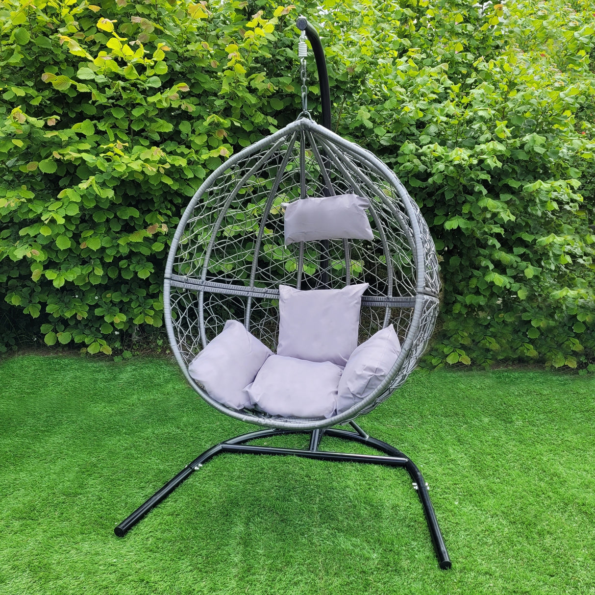 Chaise suspendue en rotin gris avec coussins moelleux, suspendue à un support noir, entourée de verdure luxuriante