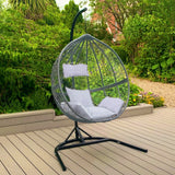 Chaise suspendue en rotin gris avec coussins blancs, suspendue sur un support noir, entourée de verdure et d'un sol en bois clair